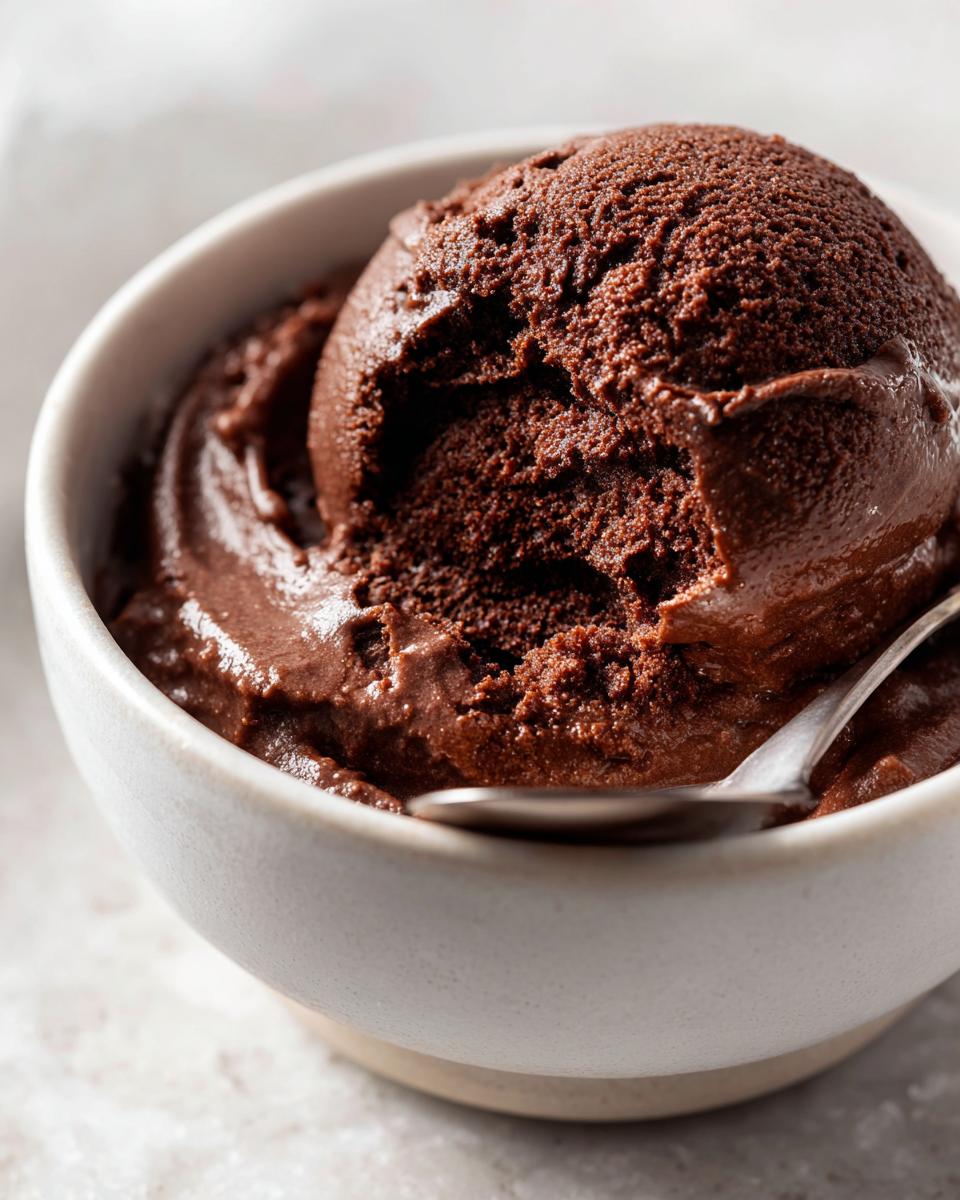 A close-up of a perfectly scooped portion of rich, dark homemade chocolate pudding in a light ceramic bowl with a spoon.