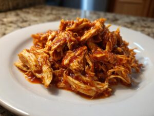 A close-up shot of shredded chicken tinga coated in a vibrant, savory red sauce piled on a white plate.