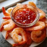 A plate of perfectly cooked shrimp surrounding a small glass bowl of bright red, chunky dipping sauce for shrimp cocktail.
