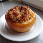 A single, glistening baked apples dessert served on a small white plate with a crispy, spiced crumble filling.