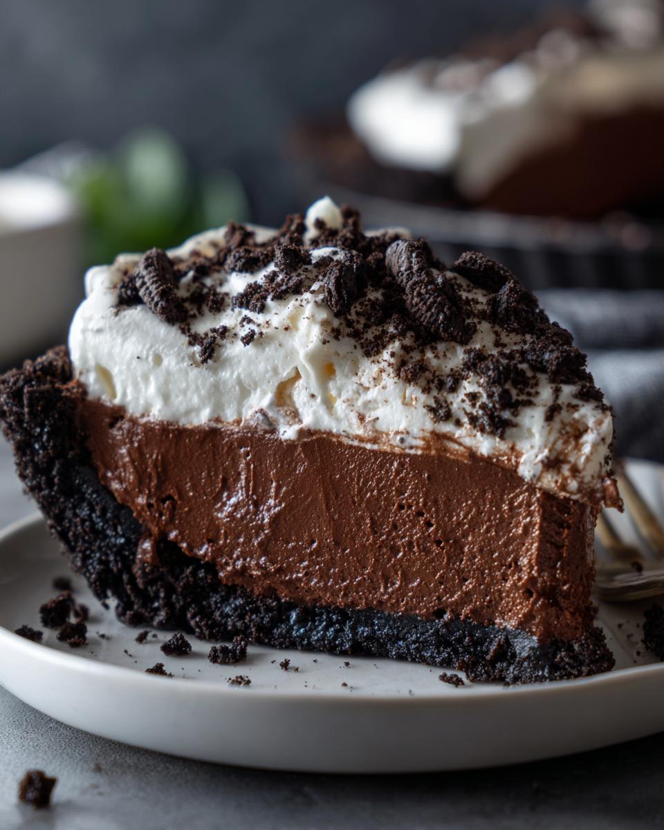 A close-up of a rich slice of chocolate mousse pie with a dark crust, topped with whipped cream and cookie crumbs.