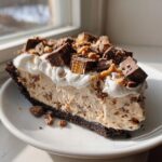 A generous slice of rich, creamy butterfinger pie with a dark crust, topped with whipped cream and chopped Butterfinger pieces.