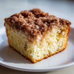A close-up of a moist slice of coffee cake featuring a thick, cinnamon streusel topping on a white plate.