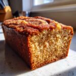A close-up, sunlit view of a freshly baked snickerdoodle banana bread loaf showing the crumb texture and cinnamon sugar topping.