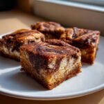 Four square snickerdoodle blondies with a cinnamon swirl topping served on a white plate.