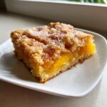 A square slice of southern peach cobbler with a thick, gooey peach filling and sugary crumb topping on a white plate.