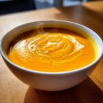 A close-up of a steaming bowl of vibrant orange squash soup, topped with a swirl of cream and cracked black pepper.