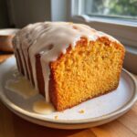 A vibrant orange sweet potato pound cake loaf topped with thick white glaze, sitting on a speckled plate.