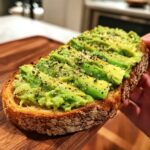 Close-up of a thick slice of avocado toast, topped with mashed avocado and cracked black pepper.