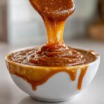 Close-up of thick, rich caramel sauce being poured from a spoon into a small white bowl.