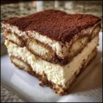 Close-up of a layered slice of tiramisu cheesecake, featuring ladyfingers, cream, and a rich cocoa powder topping.