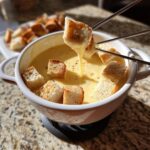 Close-up of toasted bread cubes being dipped into a white pot of smooth, yellow cheese fondue using fondue forks.