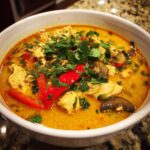 Close-up of a rich, yellow coconut curry soup filled with chicken pieces, red bell peppers, mushrooms, and fresh cilantro.