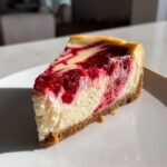 A perfect slice of white chocolate raspberry cheesecake showing a rich raspberry swirl on a white plate.
