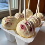 Several white chocolate covered red velvet cake pops with red sprinkles resting in a white holder.