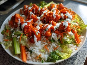 A vibrant buffalo chicken bowl featuring bright orange sauced chicken pieces over rice, lettuce, carrots, celery, and drizzled with ranch dressing.