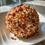 A perfectly formed cheese ball recipe coated entirely in chopped pecans, sitting on a white plate.