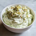 A close-up of a fluffy, light green pickle dip topped with chopped pistachios and dill in a white bowl.