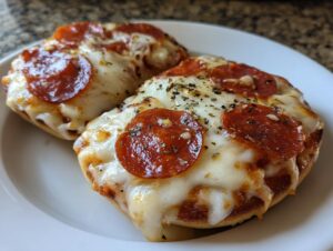 Close-up of two freshly baked pepperoni pizza bagels topped with melted mozzarella and herbs on a white plate.