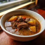 Close-up of a white bowl filled with amazing pork stew featuring shredded meat and large chunks of potato.