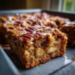 A moist square of apple brownies topped with pecans and a white icing drizzle, showing chunks of apple inside.