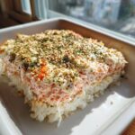 A square portion of baked salmon sushi bake casserole showing layers of rice, creamy salmon topping, and sesame seeds.