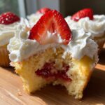 Close-up of a strawberry shortcake cupcake with a bite taken out, revealing strawberry filling, whipped cream, and a fresh strawberry slice.