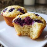 A close-up of two blueberry protein muffins on a white plate, one having a bite taken out showing the soft, yellow interior.