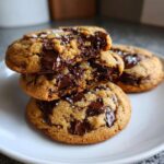 A stack of three gooey brown butter chocolate chip cookies, one broken open to show melted chocolate and sea salt.