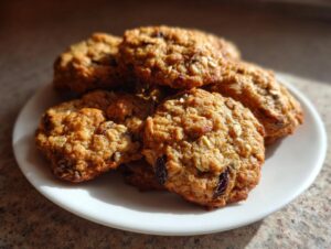 A close-up of several golden brown oatmeal raisin cookies stacked on a white plate, catching warm sunlight.