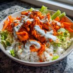 A close-up of a vibrant buffalo chicken bowl featuring orange sauced chicken, white rice, shredded lettuce, carrots, celery, and a ranch drizzle.