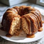 A moist apple bundt cake, drizzled with caramel sauce, cut in half to show the apple chunks inside.