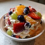 A close-up of a bowl filled with creamy cheesecake fruit salad topped with strawberries, blueberries, grapes, and orange segments.