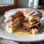 A tall stack of fluffy cinnamon roll pancakes cut open, generously topped with white icing and syrup.
