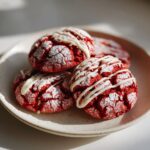 A stack of vibrant red velvet cookies, dusted with powdered sugar and drizzled with white chocolate.