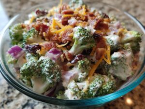 Close-up of a creamy broccoli salad featuring fresh broccoli florets, red onion, dried cranberries, bacon bits, and shredded cheese.