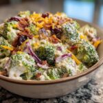 A close-up of creamy broccoli salad topped with bacon, shredded cheese, dried cranberries, and nuts.