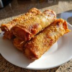 Close-up of three golden brown, crispy homemade egg roll appetizers stacked on a white plate.