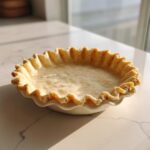 A perfectly formed, unbaked butter pie crust with crimped edges sitting in a white dish.