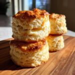 A close-up stack of four tall, flaky buttermilk biscuits with golden-brown tops resting on a wooden cutting board.