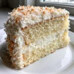 Close-up of a moist slice of fluffy coconut cake layered with white frosting and topped with toasted coconut flakes.