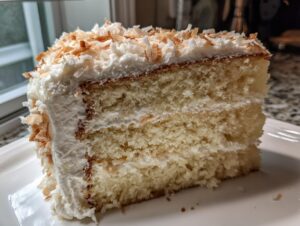 Close-up of a moist slice of fluffy coconut cake layered with frosting and topped with toasted coconut flakes.