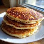 A close-up stack of three golden-brown sourdough pancakes with visible air pockets, sitting on a white plate.