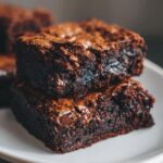 Two stacked, incredibly fudgy brownies with shiny, cracked tops and visible melted chocolate chips.