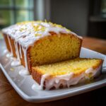 A moist, yellow lemon loaf drizzled with white icing and topped with lemon zest, with one slice cut and resting beside it.