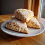 A stack of three soft lemon scones topped with white glaze and lemon zest, resting on a white plate.