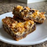 Two stacked, gooey magic cookie bars featuring chocolate chips and toasted coconut topping on a white plate.