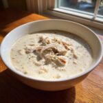 A close-up of a white bowl filled with rich homemade cream of chicken soup, topped with shredded chicken and herbs.