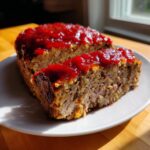 A thick slice of juicy meatloaf topped with a shiny, red glaze, sitting next to the rest of the loaf on a white plate.