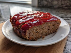 A thick slice of juicy meatloaf topped with a glossy, bright red glaze, served on a white plate.